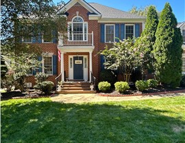 Front view of a red brick two-story house with a new Charcoal Timberline HDZ roof, white porch railing, blue door, matching shutters, and a vibrant green lawn under a clear blue sky in Chesterfield, VA.
