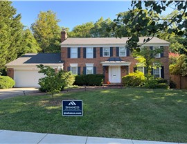Two-story brick home in Alexandria, VA, with black shutters and a newly replaced roof featuring Pewter Gray Timberline HDZ shingles. The home is set against vibrant blue skies and a lush, tree-lined backyard offering ample shade.