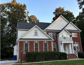 A two-story brick and white siding house with Pewter Gray Timberline HDZ shingles, a front balcony, and white railings, surrounded by a green lawn and tall trees under a clear blue sky in Midlothian, VA.
