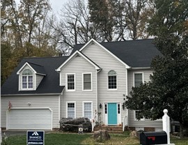 Front view of a two-story home with a new Charcoal Timberline HDZ roof, light gray siding, white trim, and a teal front door. The yard features a Shanco sign, and mature trees in the background on a cloudy day in Glen Allen, VA.