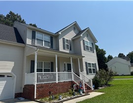 Front view of a beige two-story home with a new Charcoal Timberline HDZ roof, white front porch railing, dark green shutters, and a brick foundation. The home is situated on a well-kept lawn with landscaping, under a clear blue sky in Chesterfield, VA.