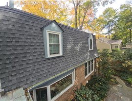 Brick house with gray-blue accents and a Mansard roof featuring Pewter Gray Timberline HDZ shingles, surrounded by mature trees and lush landscaping in Rockville, Maryland.