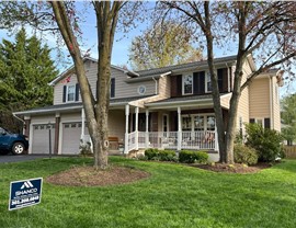 Cream-colored home in Fairfax, VA, with black shutters and brick accents, showcasing a new roof replacement with Pewter Gray Timberline HDZ shingles from GAF. The property is surrounded by large trees in both the front and backyard.