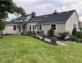 Beige home in Manassas, VA, with a newly installed Charcoal Timberline HDZ shingle roof. The house features a spacious lawn, vibrant landscaping, a stone pathway, and mature trees providing shade. The sky is slightly overcast, adding a peaceful ambiance.