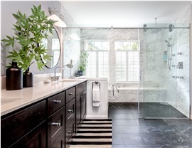 Modern bathroom remodel with dark wood vanity, marble walls, frameless glass shower, and freestanding tub beneath shuttered windows.