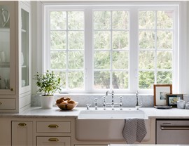 Bright farmhouse-style kitchen with apron-front sink, marble countertops, and large divided-light windows overlooking greenery.