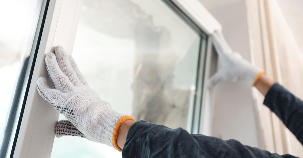 A person wearing white work gloves presses their hands against the frame of a window they're installing.