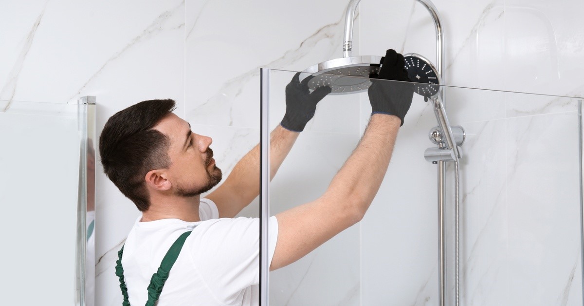 A person wearing black gloves standing inside a shower booth, with both hands placed on a showerhead.