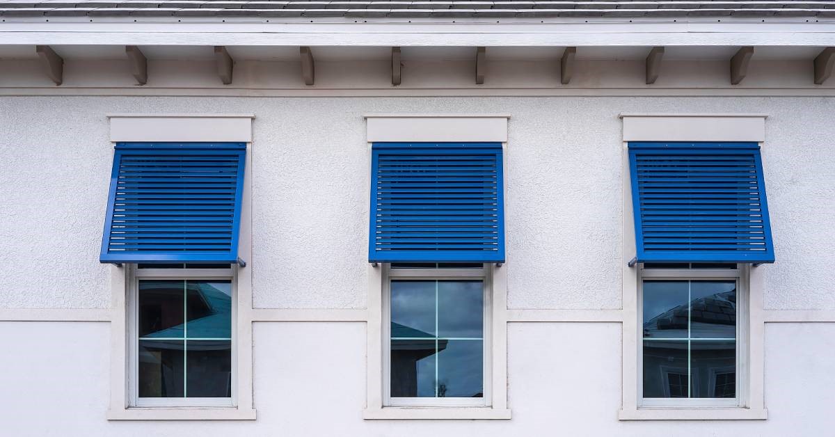 A slate-gray residential beach house boasting three identical hurricane windows with blue hurricane shutters.