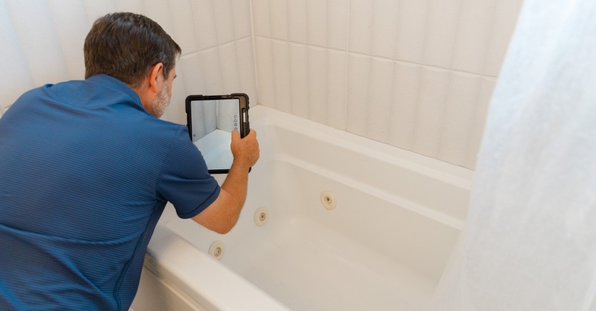 A man in a blue shirt is kneeling over a bathtub. He’s holding a tablet and examining the grout.