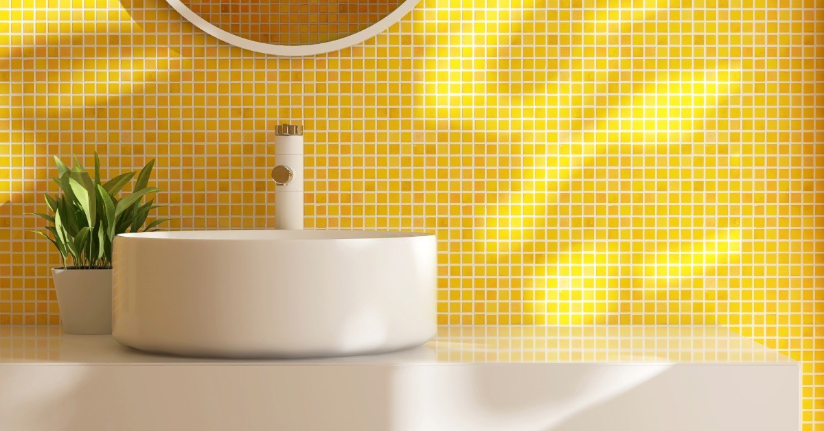 A sink with a circular bowl in installed next to a yellow tile wall. There is a circular mirror above the sink.