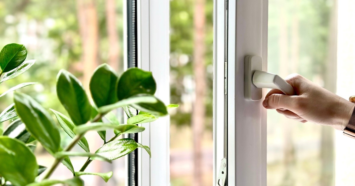 A hand is opening a casement window, with a view of greenery, sky, and trees outside.