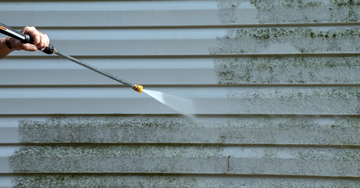 A person uses a pressure washer hose to remove a layer of mold from the vinyl siding of a property's exterior.