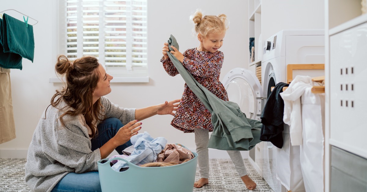 A woman in a gray sweater and a small child wearing a floral shirt are taking laundry out of a washing machine.