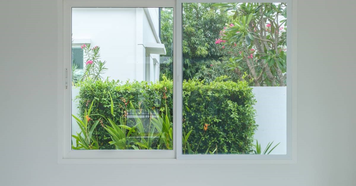 A sliding window surrounded by a white wall in a house. The window shows a view of some flowers, trees, and shrubs.