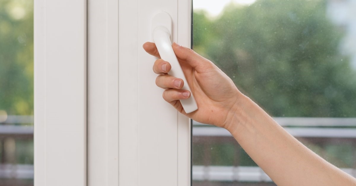 A close-up of a person using the handle on a white casement window. Blurred trees are visible beyond the window.