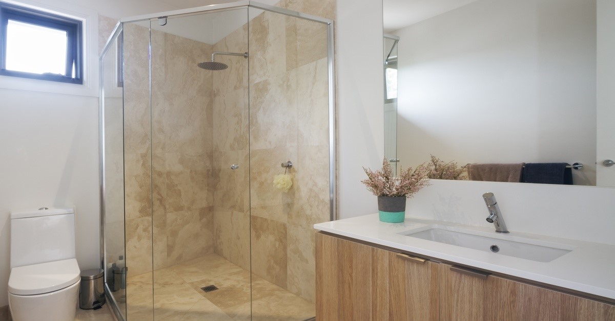 A corner shower with marble tiling installed between a white toilet and a mirror and vanity cabinet set in a bathroom.