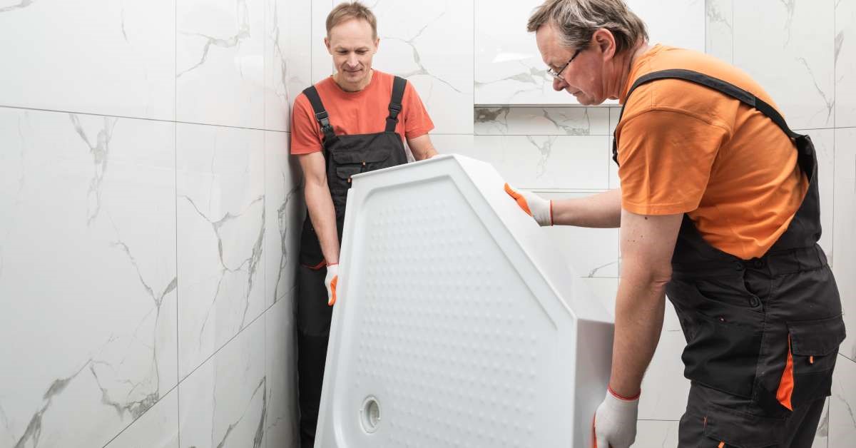 Two professional contractors removing a shower tray from the bathroom to prepare for a dual-purpose tub installation.