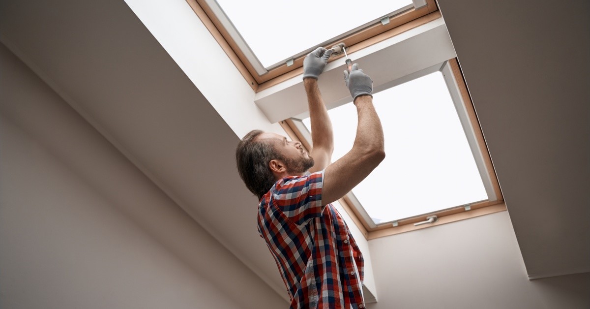 A man in a flannel shirt is using a screw driver to tighten a window handle. The window is attached to the ceiling.