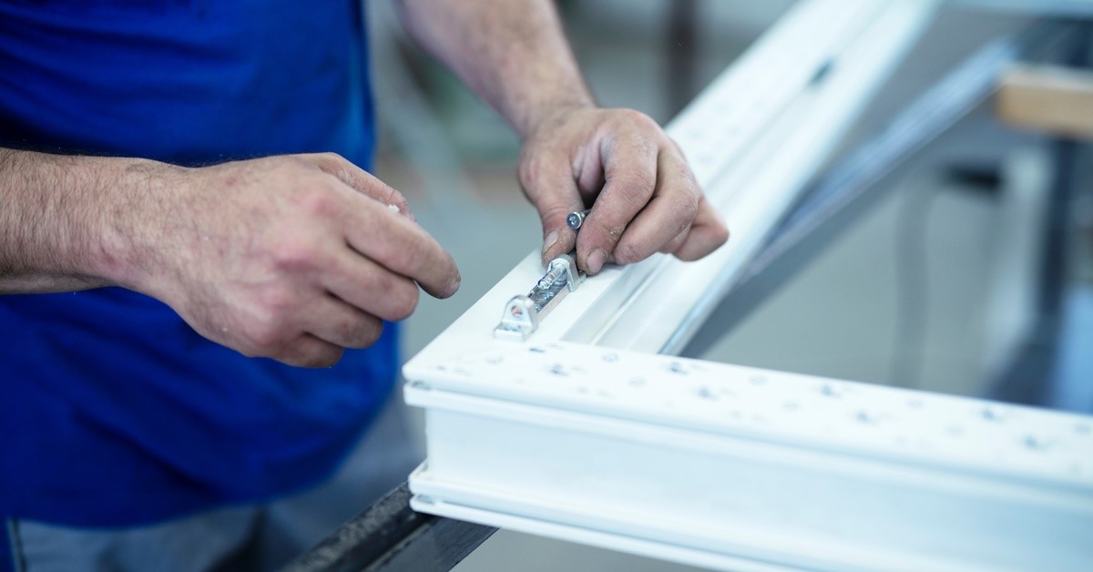 The hands of a person wearing a blue shirt as they assemble a hinge piece onto a white PVC casement window.