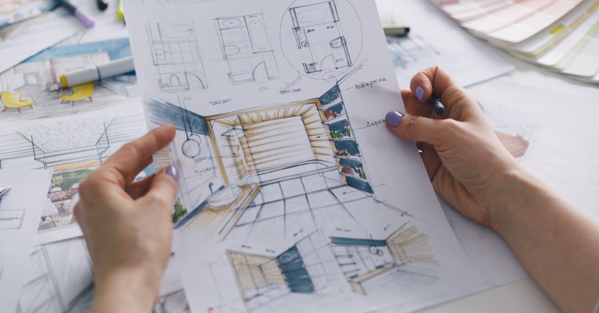 A person is holding a piece of paper with a drawing of a bathroom on it. There are other papers on the desk.
