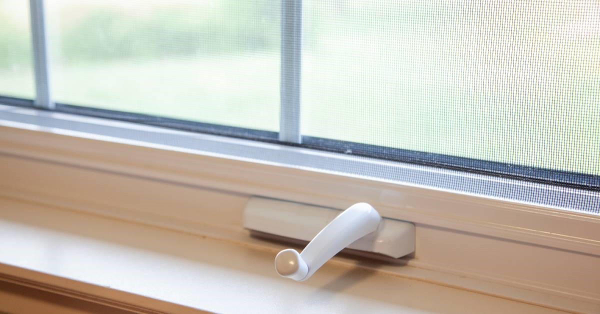 A close-up of the hand crank mechanism on a casement-style window. The window also features a screen.