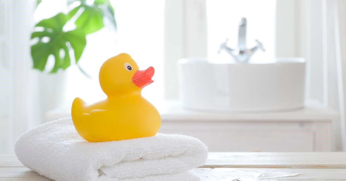 A large rubber duck bath toy resting on top of a neatly folded bath towel with a child’s bathroom in the background.