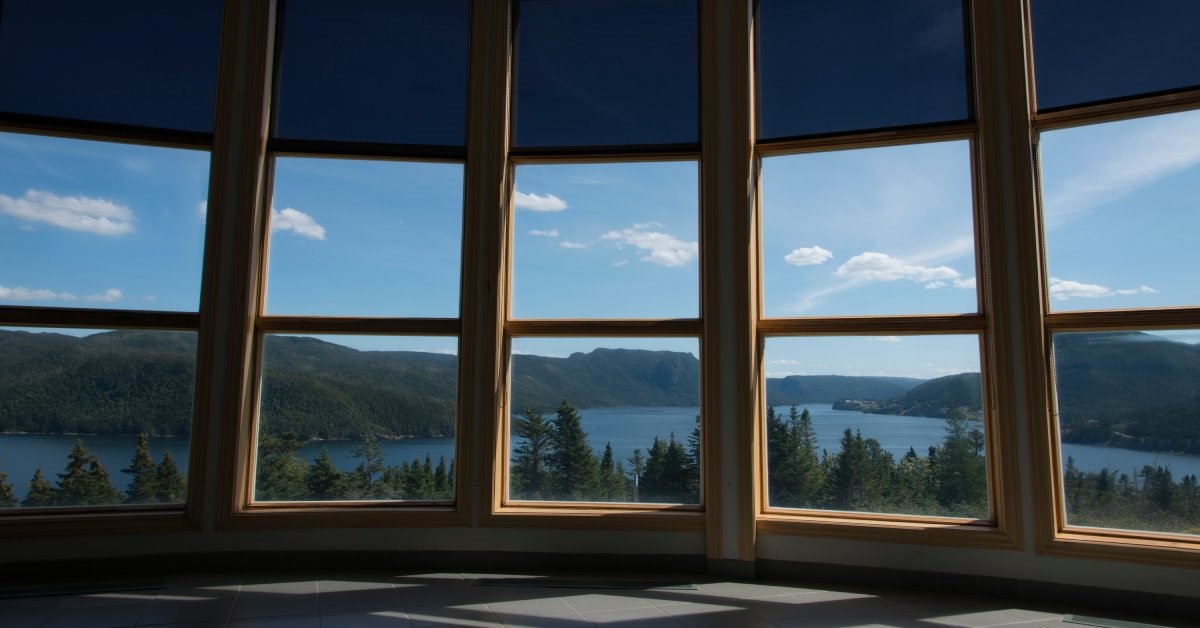 The inside of a large room with floor-to-ceiling bow windows overlooking a picturesque alpine lake and forest.