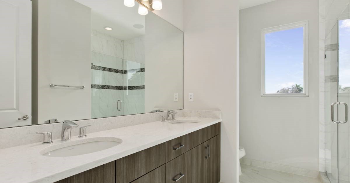 A modern home bathroom featuring white marble countertops with modern sink fixtures. There is a shower across from the vanity.