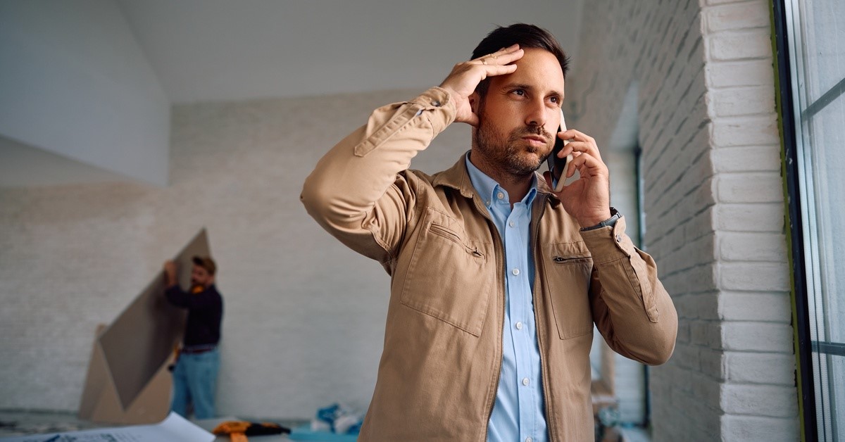 A man with short brown hair is standing in a room. He’s holding a phone up to his ear and is placing his other hand against his head.