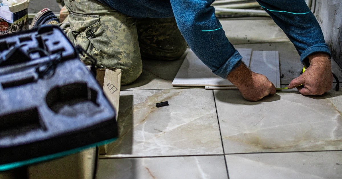 A person in a blue shirt and camo pants is laying tile in a bathroom. There is a toolbox next to them.