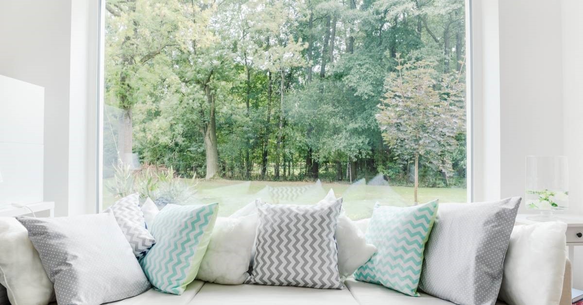 A close-up of a window seat in front of a picture window overlooking a tree-lined yard. There are many pillows lining the seat.