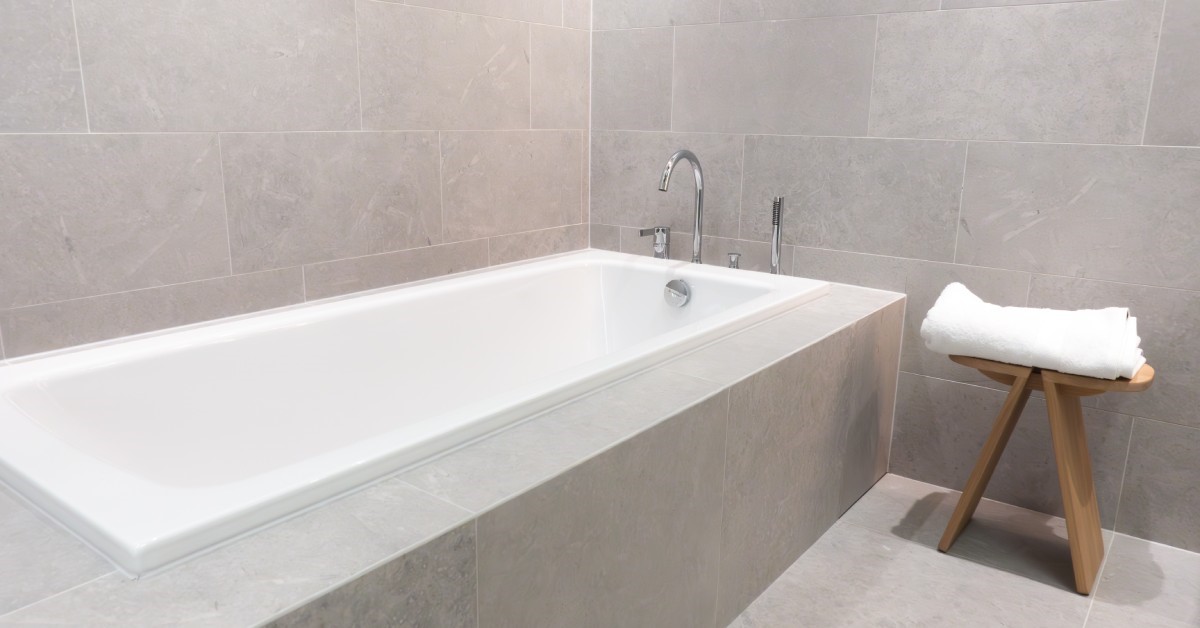 A built-in tub is attached to a wall that has gray tile. There is a stool next to the tub with a towel on it.