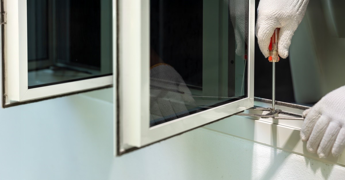 A person is holding a screwdriver and pressing it against a windowsill. The windows are open and the person is wearing gloves.