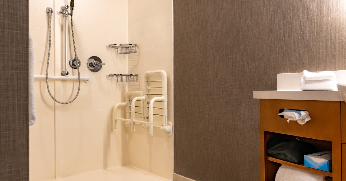 An accessible, roll-in shower stall with a zero-threshold entry, wall-mounted grab bars, a bench, and a shower faucet wand.