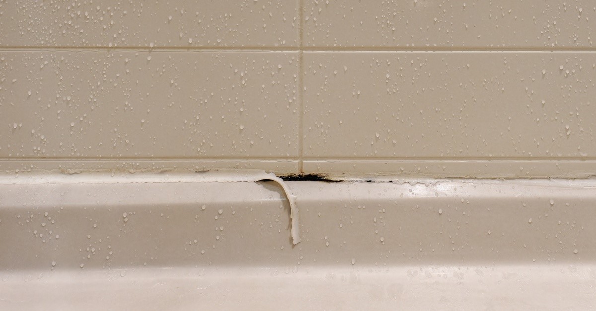 A white wall in a shower with water droplets on its surface. The caulking in the shower is peeling and mold grows underneath.