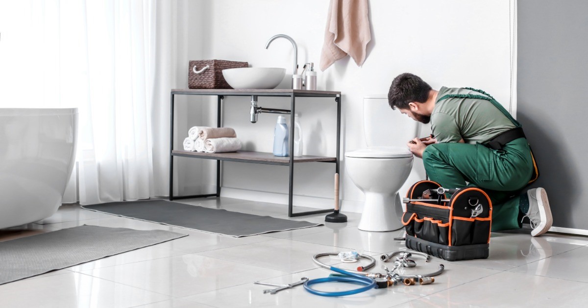A man is kneeling next to a toilet. He has a tool box next to him as he works on the toilet.