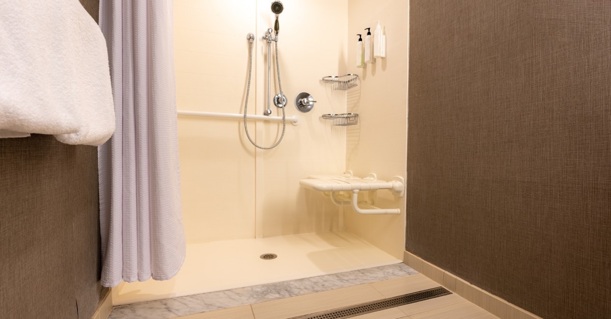 A roll-in shower with tiled floors and walls. The shower includes an adjustable showerhead and a seat mounted to the wall.