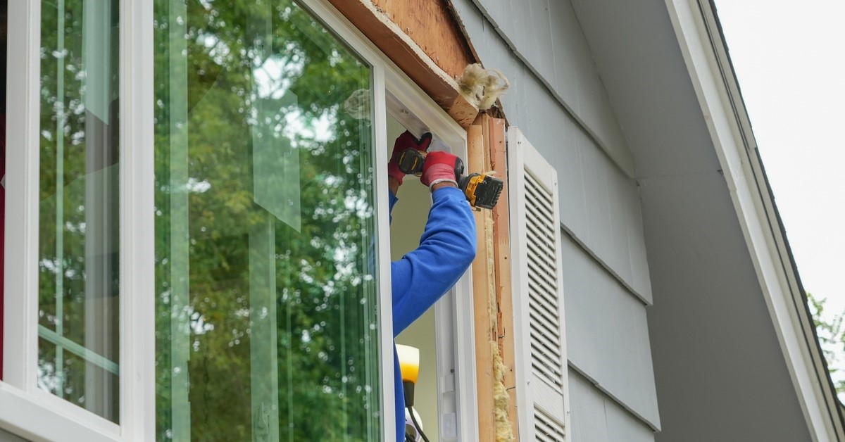 A man in a blue shirt is using a drill on a home. He’s drilling into the wood of a window frame.
