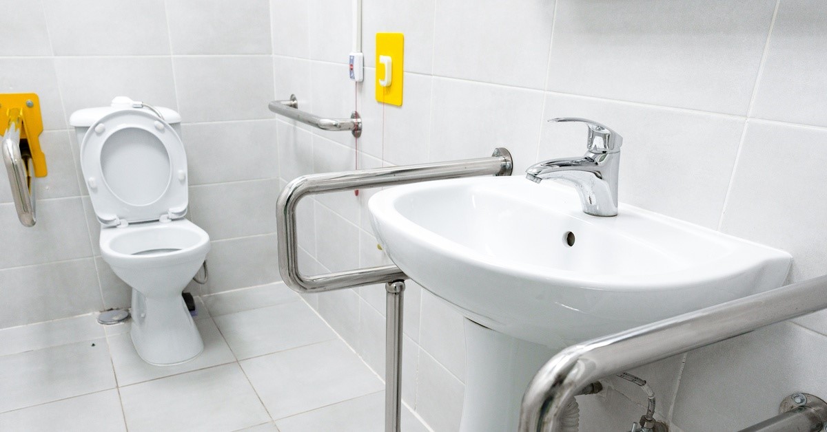 A bathroom with white tiling for the floors and walls, with safety rails near the toilet and by the sink.