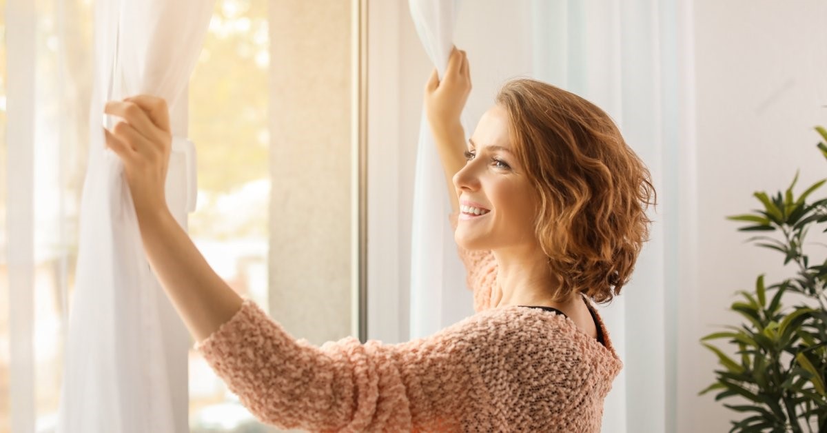 A young woman in a pink sweater opening her window curtains and smiling as natural light floods her home's interior.