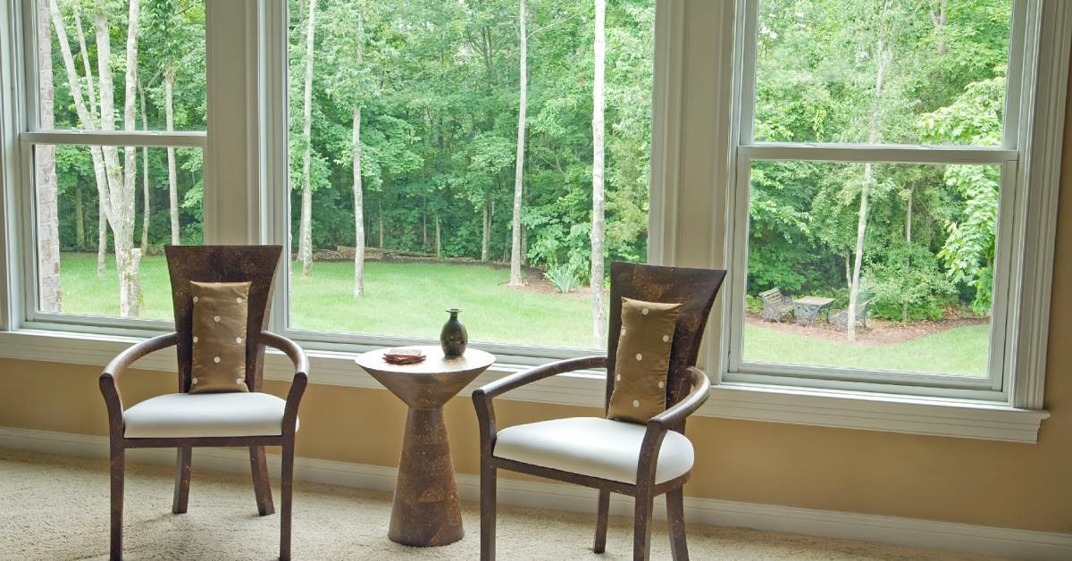 A sitting area with two chairs and a small table set up in front of a large window. Many trees are visible through the window.