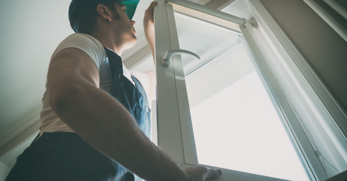 Light shines through the window glass as a person wearing work clothes installs the window frame in place.