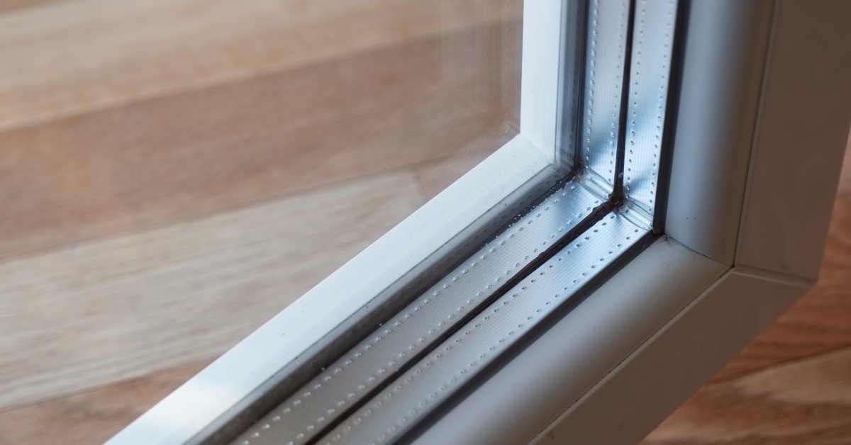 A close-up of a glass window with triple glazing and a neutral colored frame. The glass reflects a wooden surface.
