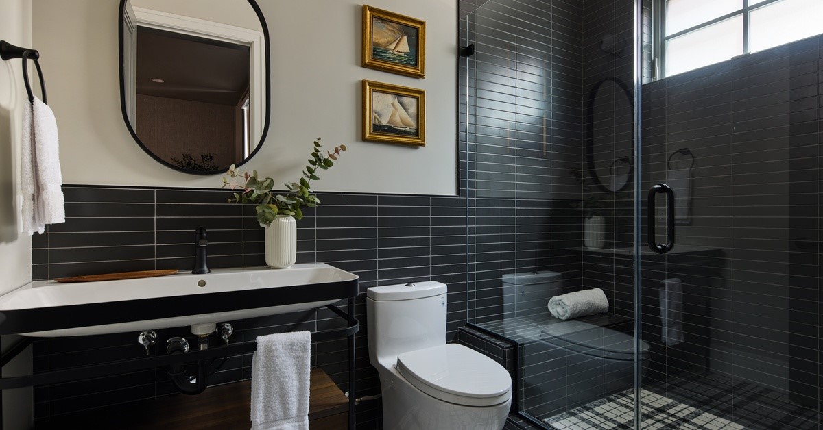 A bathroom with black tile running along the bottom third of the wall. There is a window in the shower.