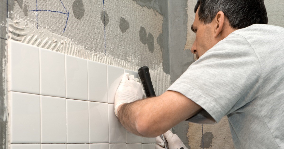 A man is fitting tiles into a wall. He is using the handle of a hammer to place the tiles onto the wall without damaging them.