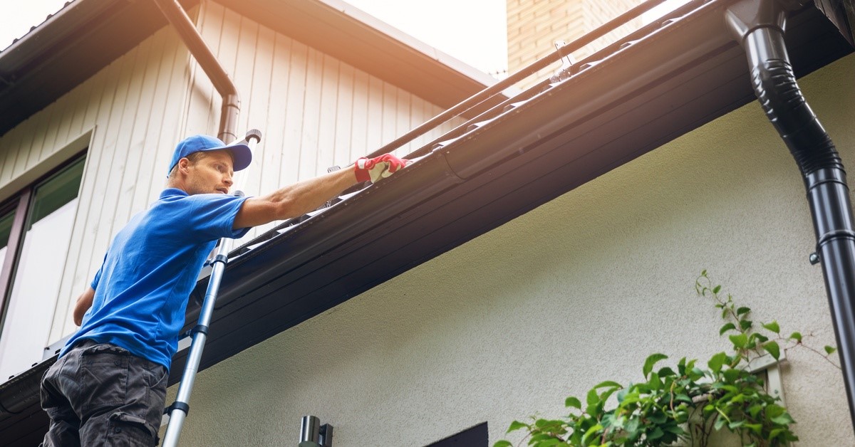 A man in a blue shirt and hat is reaching into a gutter of a home. There is a chimney in the background.