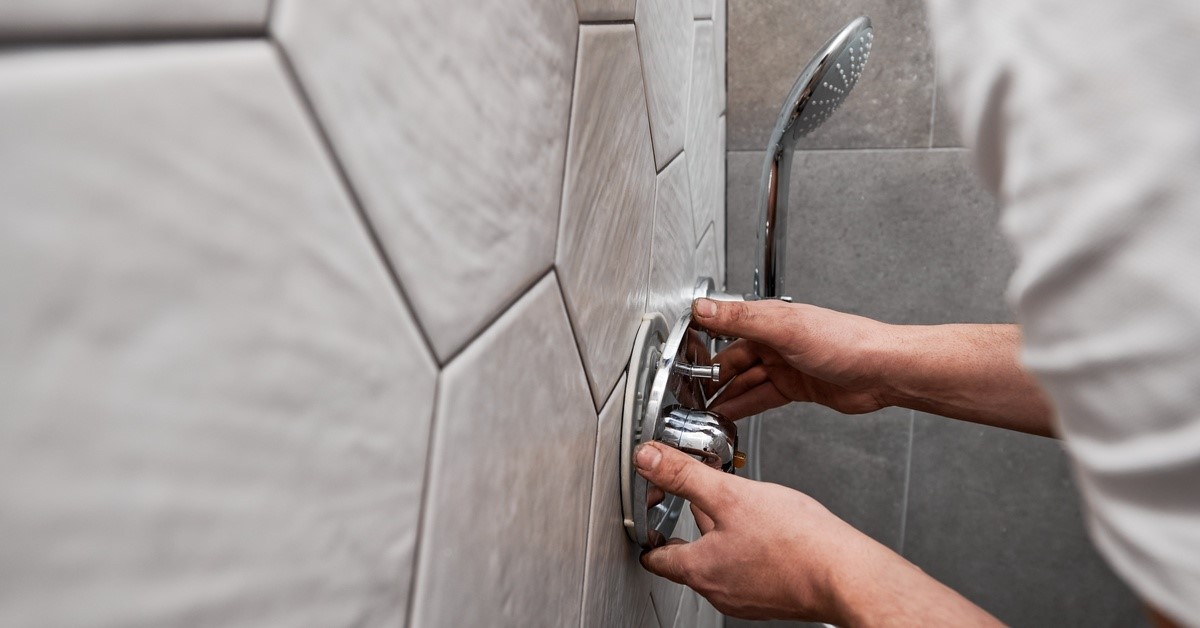 A person's hands as they install a shower faucet with a metallic handle onto the ceramic tile of the shower wall.