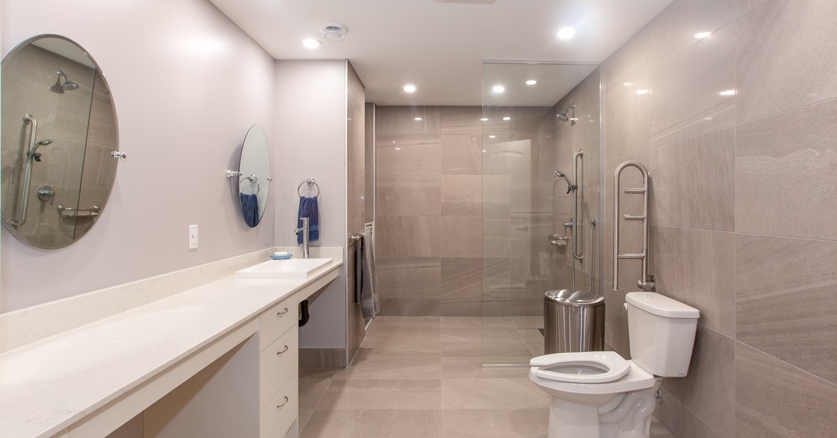 A bathroom with gray tile flooring and a silver metal handle next to the toilet. There are two mirrors.