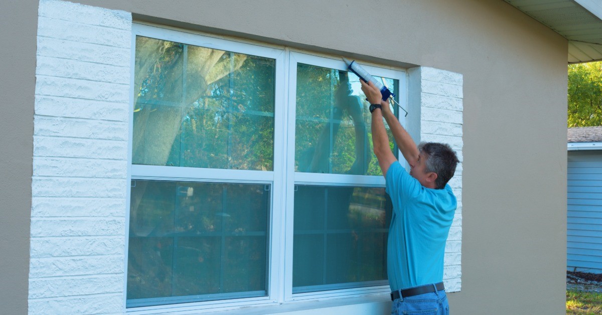 A man is using a calk gun to seal a window from the exterior. There are shutters on either side of the window.